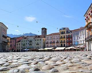 Vista di una piazza con edifici storici e montagne sullo sfondo.